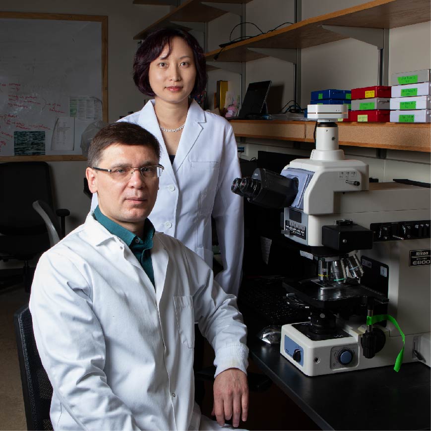 Qixin He and Pavel Klimov wearing lab coats. He standing and Klimov sitting beside a lab bench